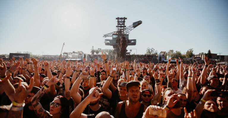 A large crowd of people at an outdoor music festival, many with their hands raised in the air, with a stage structure visible in the background under a clear sky.