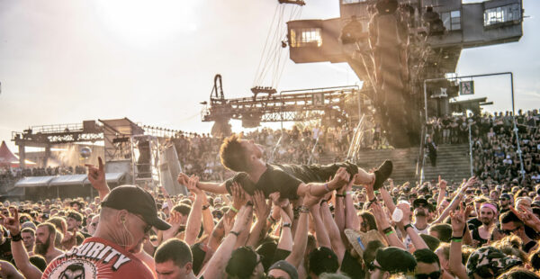 Crowd at an outdoor music festival with a person crowd surfing above a cheering audience, with a large stage structure and setting sun in the background.