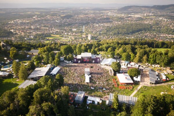 Aerial view of an outdoor music festival with stages and a large crowd of attendees, set among green trees with a panoramic backdrop of a cityscape.