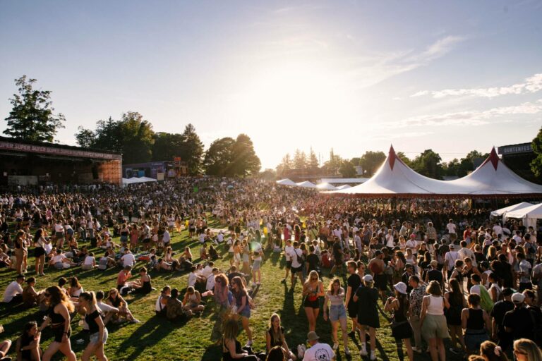 A large crowd of people enjoying an outdoor music festival with tents and a stage visible, under a bright sunlit sky.