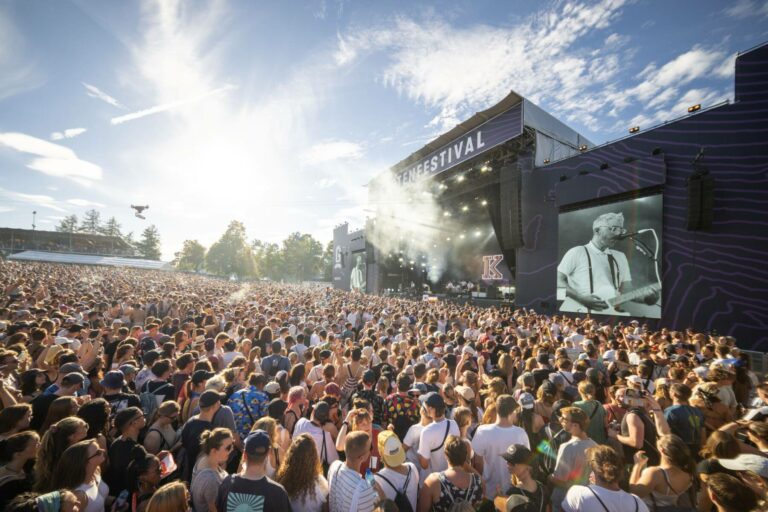 A large crowd of attendees enjoying an outdoor music festival in the sunlight, with the stage in the background displaying a live performance on a big screen.