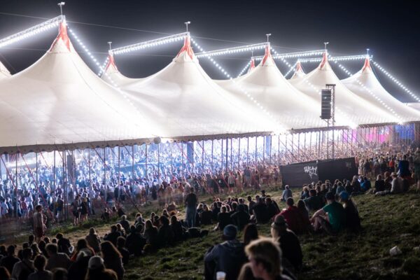 Large festival tent illuminated by strings of lights at night with crowds of people inside and sitting on a hillside in the foreground.