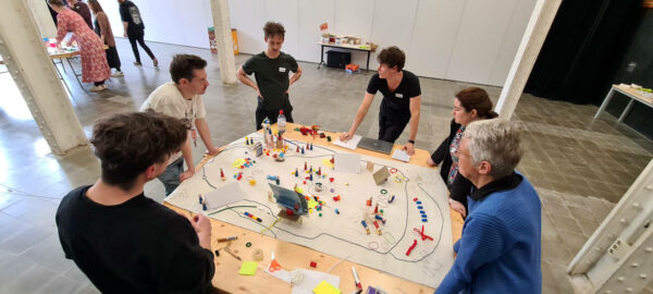 Group of people standing around a table with various colorful objects and materials on it, apparently engaged in a collaborative activity or workshop.
