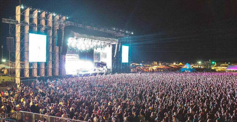 A large outdoor music festival at night, featuring a crowded audience in front of a brightly lit stage with large screens on either side.