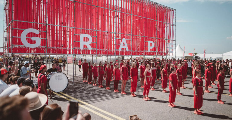 Group of people dressed in red arranged in lines before a large red hanging ribbon installation with the letters 