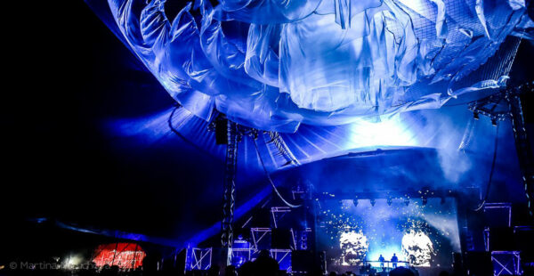 A concert stage illuminated by blue lights with a large, undulating inflatable object overhead, and a crowd silhouetted at the bottom in front of a video screen displaying abstract visuals.