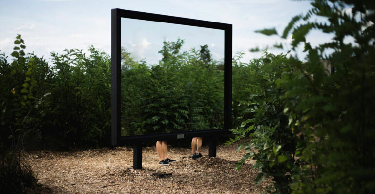 A large, freestanding picture frame outdoors with the lower half of a person's body visible through it, creating the illusion of the person blending into the surrounding greenery.