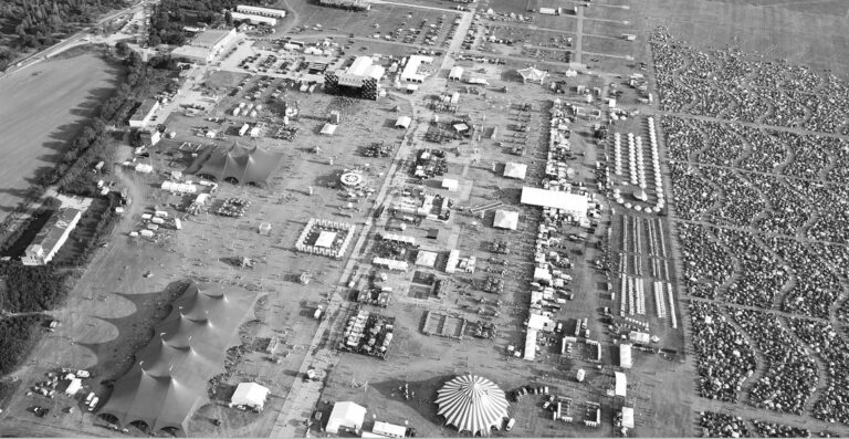 Aerial black and white photograph of an extensive outdoor festival area with multiple large tents, stages, rows of parked cars, and surrounding campgrounds.