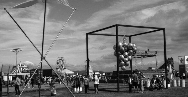 Black and white photo of an outdoor festival scene with people, a Ferris wheel in the background, a tent to the left, and a structure with hanging mirrored disco balls in the foreground.