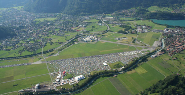 Aerial view of a large outdoor event with numerous tents and a massive gathering of people in a green valley, adjacent to a small town and a lake.