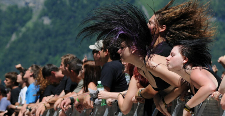 A crowd of enthusiastic concertgoers with a few individuals headbanging, their hair wildly in motion, in front of a metal barrier with a green mountainous backdrop.