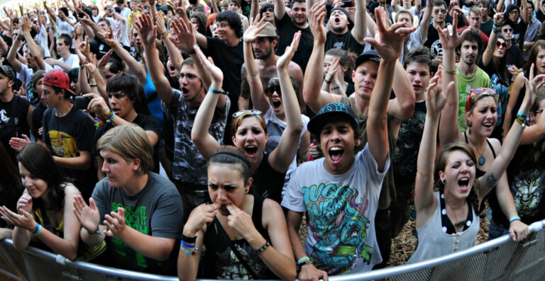 A crowd of excited concertgoers cheering and clapping, some with arms raised, at an outdoor music festival.
