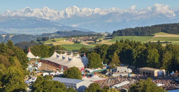 A panoramic view of an outdoor festival with large tents and gathered crowds, set against a backdrop of rolling hills and distant snow-capped mountains.