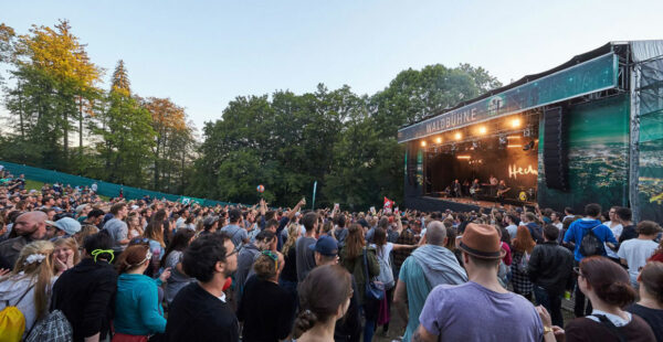 Crowd of people enjoying a live concert at an outdoor venue surrounded by trees, with the stage displaying the word 