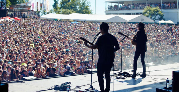 Two musicians are seen from behind performing on stage in front of a large, densely packed audience at an outdoor concert venue during the daytime.