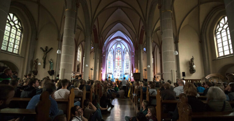 A congregation of people sitting inside a cathedral with tall arched ceilings and stained glass windows, focusing on a performance near the altar.