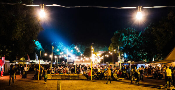 Nighttime scene at an outdoor festival with illuminated tents, crowds of people mingling, and several bright lights hanging overhead.