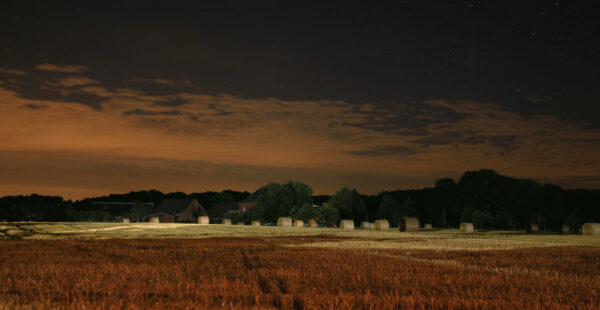 A nighttime scene of a harvested field with round hay bales, farm buildings in the background under a sky with orange clouds and visible stars.