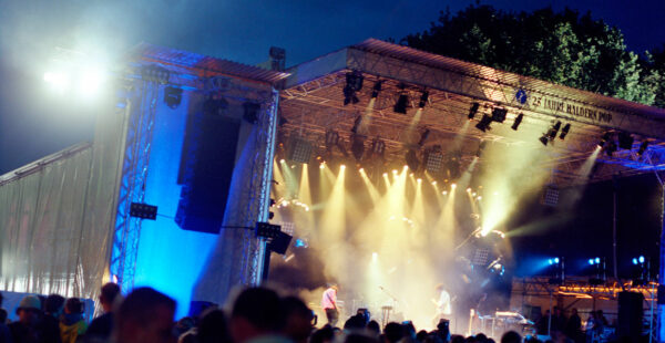 Outdoor music concert at night with bright stage lights and a band performing in front of an audience, with a banner reading 