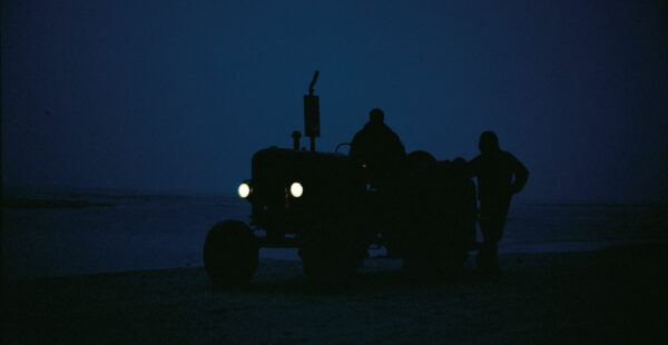 A silhouette of a tractor with its headlights on and two people, one seated on the tractor and one standing beside it, against a twilight sky.