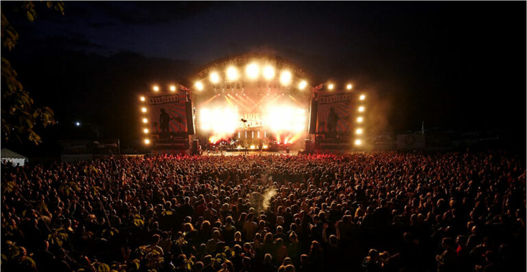 A crowded outdoor concert at night with a brightly lit stage and numerous spectators under a dark sky.