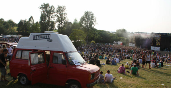Outdoor music festival scene with a large crowd sitting on the grass in front of a stage with a red van advertising 'Happiness Festival' in the foreground.