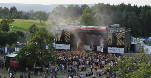A music festival scene with a crowd of people in front of a stage with large banners, surrounded by trees, and haze above the crowd, suggestive of outdoor live performance ambiance.