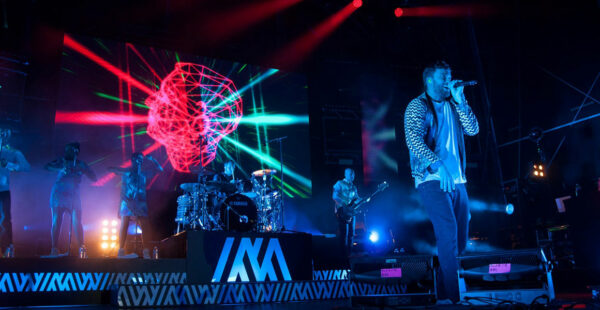A concert stage with a band performing at night, featuring colorful laser lights and visual effects. The lead singer is center stage with a microphone, flanked by background vocalists to the left and band members playing drums and guitars to the right. The stage is adorned with the band's name in large letters.