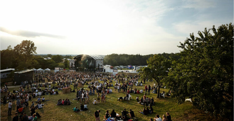 An outdoor music festival scene with a crowd of people sitting and standing on grassy hills, a stage in the background, and trees and clear sky overhead.