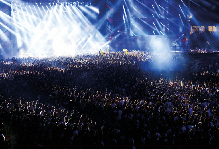 A large crowd of people at an outdoor music festival, with bright stage lights shining above them.