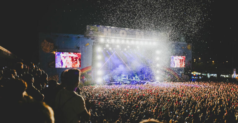 A crowd of people at an outdoor night concert with bright stage lights, large screens showing performers, and confetti falling in the foreground.