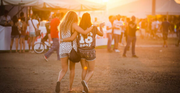 Two women with arms around each other walking at a festival during sunset, with other festival-goers and tents in the background.