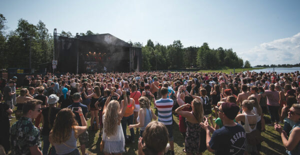 A large crowd of people enjoying an outdoor music festival by a lake with a clear sky above.