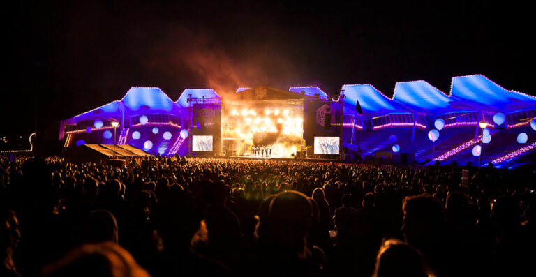 A large crowd at an outdoor night concert with a brightly lit stage and dynamic blue lighting on the stage structures.