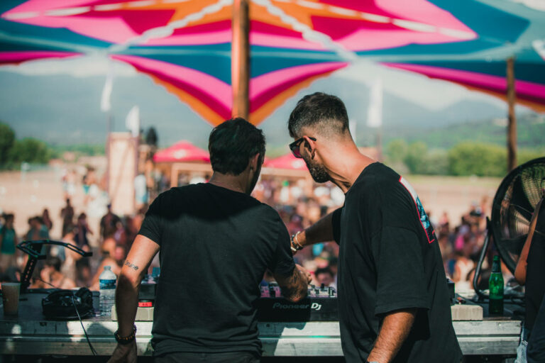 Two DJs are standing at a mixing console at an outdoor music festival with colorful canopies above and a crowd in the background.