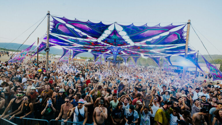 A vibrant outdoor music festival with a large crowd of people gathered under a colorful, psychedelic canopy, with hills in the background.