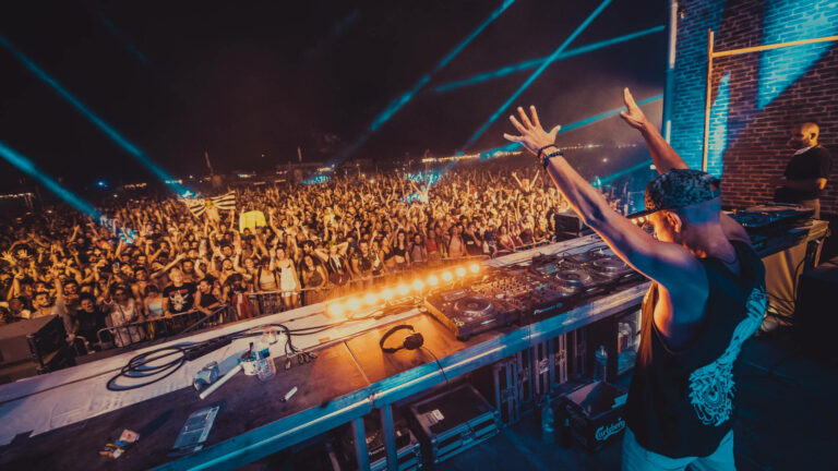 DJ performing at a music festival with his hands raised towards a large crowd under blue stage lights.