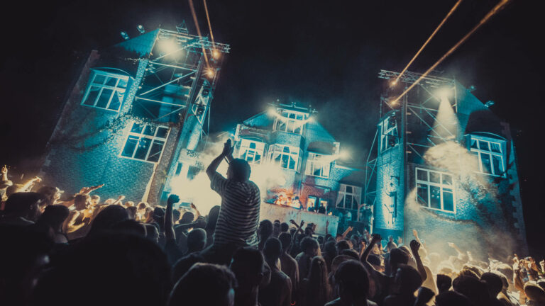 A vibrant outdoor night concert with a crowd of people cheering and raising their hands towards a lit-up stage set in front of a brick building with spotlights and beams of light crossing the sky.