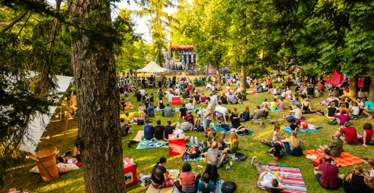Outdoor summer festival scene with people sitting on blankets and enjoying a live music performance on a stage surrounded by trees.
