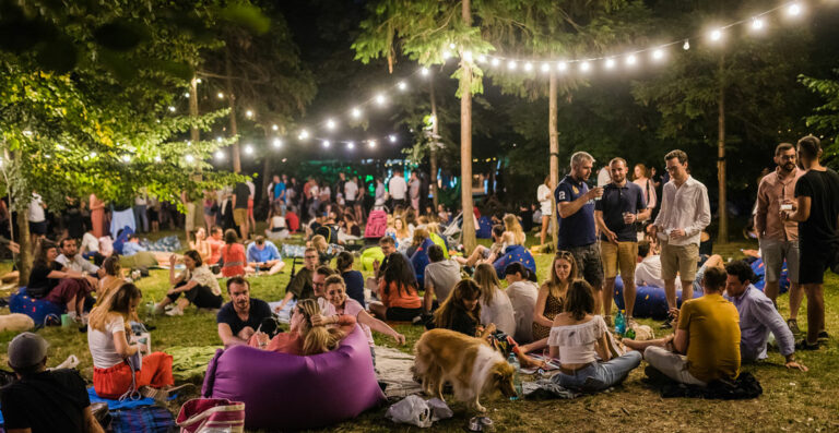 Outdoor evening gathering in a park with people sitting on blankets and inflatable loungers, some standing and talking, with string lights overhead.