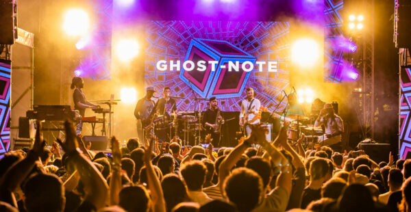 A live concert performance by the band Ghost-Note with members playing keyboards, saxophones, guitar, and drums on stage, with a vibrant backdrop displaying the band's name, surrounded by an energetic crowd cheering and raising their hands.