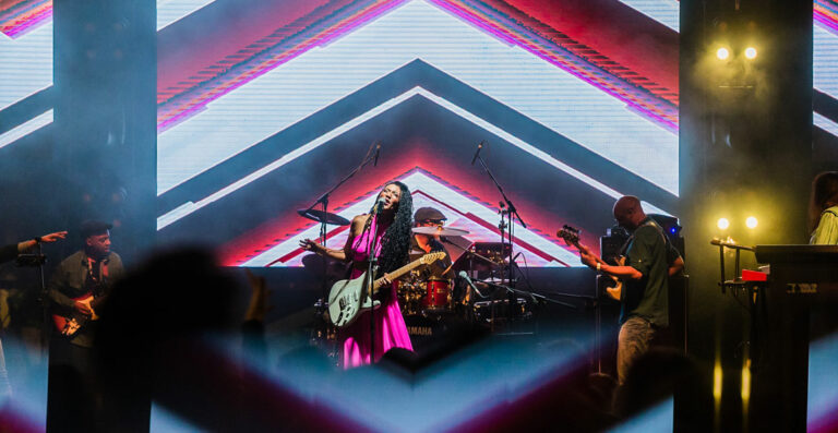 A band performing on stage with vibrant lighting and a large LED screen displaying geometric patterns in the background; the silhouette of the audience is visible in the foreground.