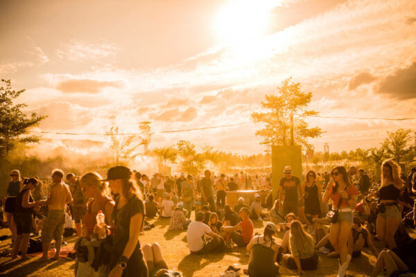 An outdoor music festival scene with people walking, sitting, and socializing in a dusty area, backlit by a soft golden sunset that gives the image a warm glow.