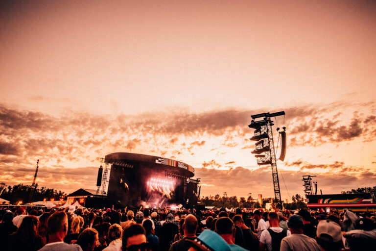 Outdoor music festival at sunset with a large crowd facing a stage with bright lights and a cloudy sky in the background.