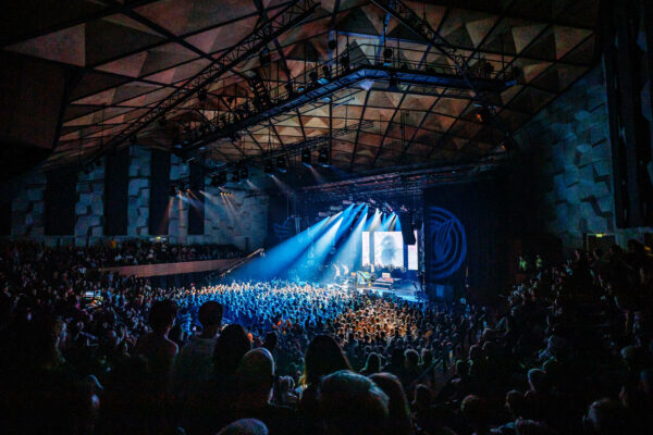 An indoor concert with a large audience watching a band perform on a stage illuminated by blue lights, with images displayed on a screen in the background.