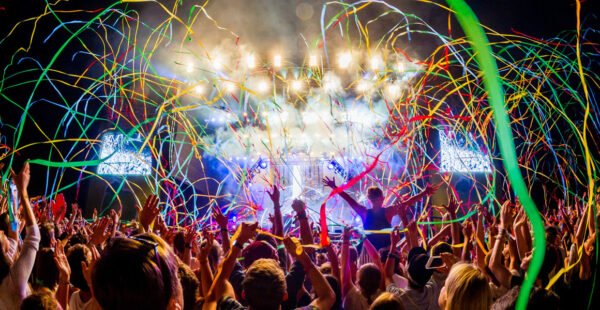 A vibrant concert crowd with raised hands, colorful streamers flying through the air, and a bright stage with lights and smoke effects in the background.