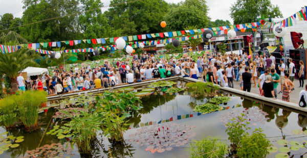 A lively outdoor party with a large crowd of people gathered during daylight, festooned with colorful flags, large white balloons, and a stage set up for a performance by a water lily pond in the foreground.