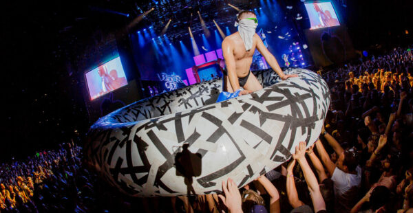 A person in underwear and a blindfold is stage diving onto an inflatable raft held by a crowd at a concert, with large screens displaying performers in the background.