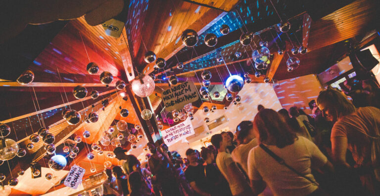 A lively indoor scene at a night club with multiple disco balls hanging from the ceiling, colorful lighting, and a crowd of people socializing and dancing.