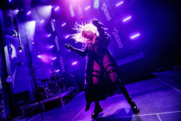 July Jones with blonde hair headbanging on stage, wearing a black outfit with fishnet tights, under purple stage lights with the text 